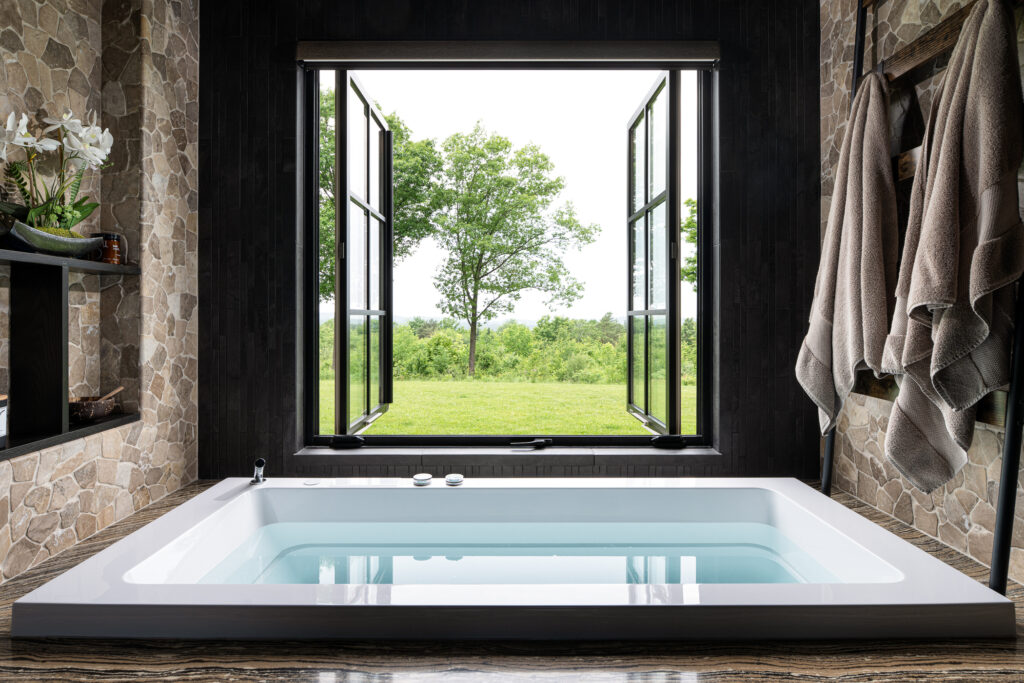 Ceramic bathtub filled surrounded by stone walls. Above the bathtub, windowpane doors open up to a lush green countryside yard in Vermont. Revealing construction details from Silver Maple.