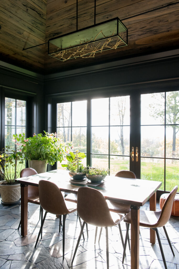 A scouting image of an interior dining space looking with afternoon light illuminating a dining table shining through floor to ceiling window paneled doors surrounded by plants.