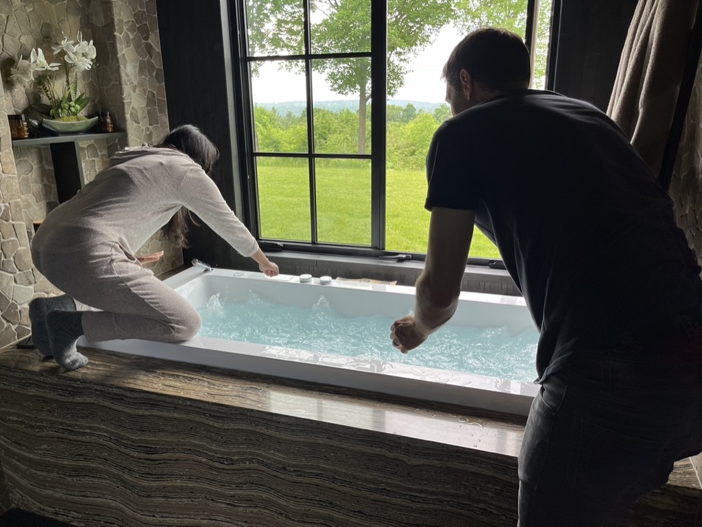 A woman kneeling over a stone bathtub filling it up assisted by a professional photographer to capture the behind the scenes photography process of capturing construction details.