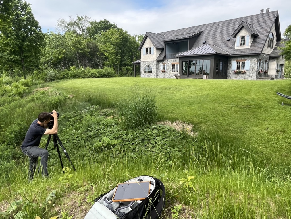 A photographer standing in a lush green lawn, pointing at a whimsical modern stone house. Showing the behind-the-scenes process of capturing the perfect photo. 