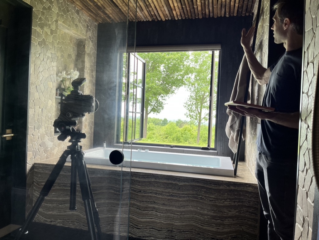 A behind the scenes look at a professional photographer, Ryan Bent, setting up his camera to capture the construction details by Silver Maple. The camera is pointing toward a stone bathtub underneath an open windowpane to a lush green countryside in Vermont.